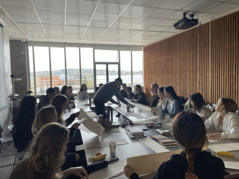 People gathered around long table on both sides, sitting and drawing while instructor is in the middle