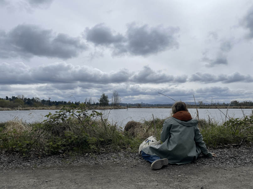 young person in blue jacket sits on ground looking out on the water