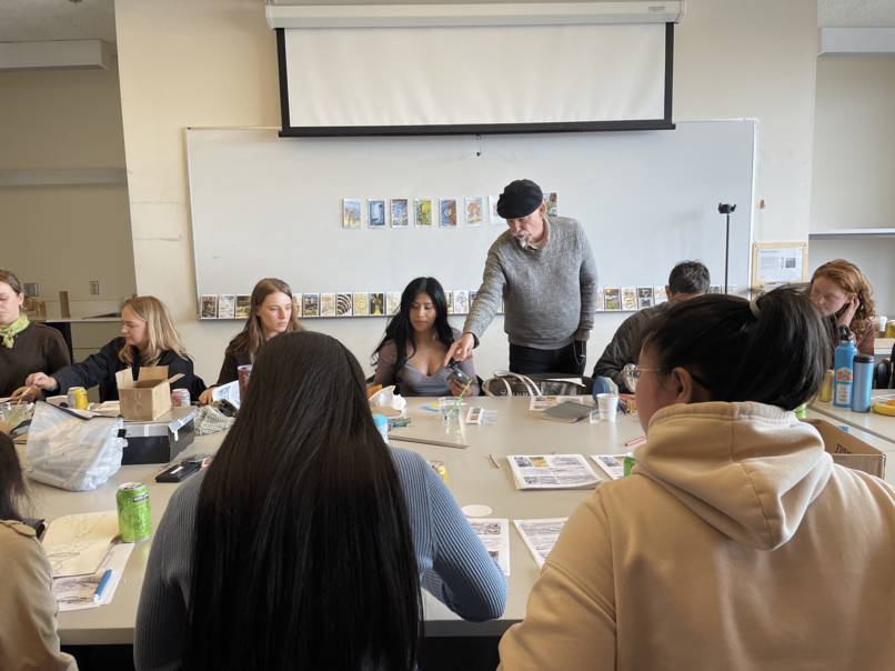 instructor in black beret points to students drawing, he is standing and students are sitting at a table